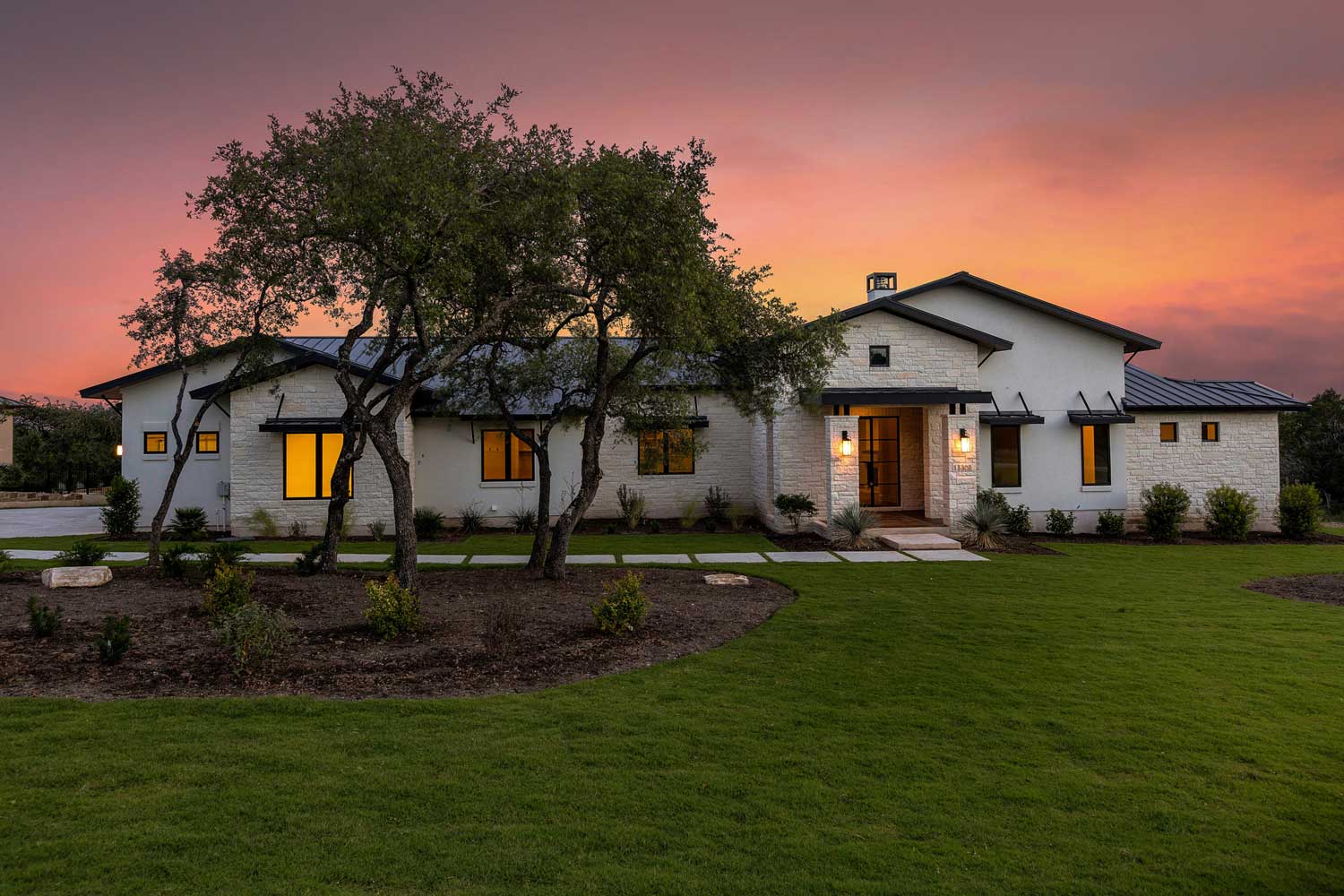 A stone-and-stucco Texas Hill Country home at dusk, framed by mature oak trees with warm light in the windows